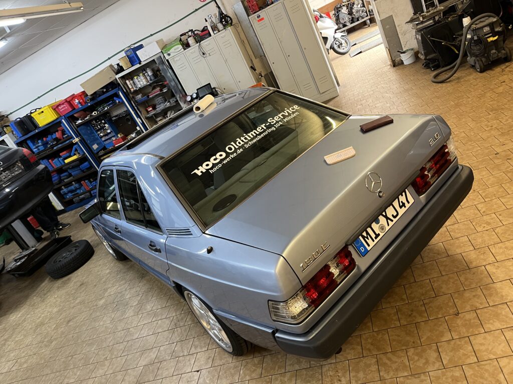 Silberner Mercedes 190E 2.0 in der Werkstatt von HOCO-Werke Minden mit Heckaufkleber „HOCO Oldtimer-Service“ – gepflegter Youngtimer auf glänzenden Felgen, fotografiert in der Werkstattumgebung mit Werkzeugschränken und Reifen im Hintergrund.