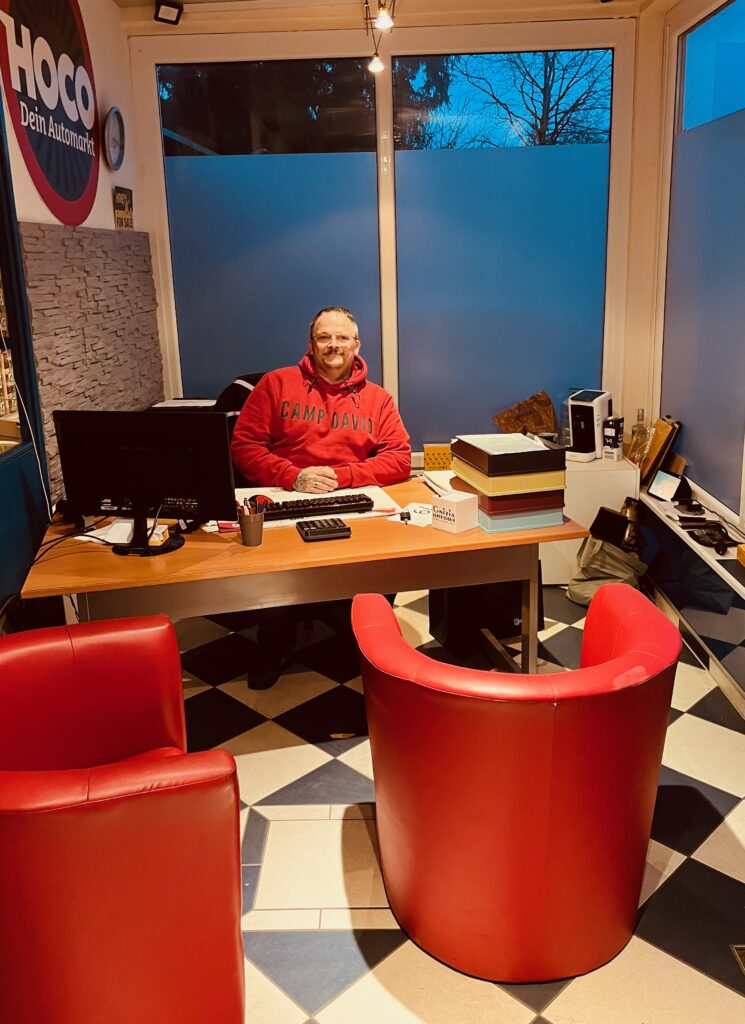 Büro des HOCO Automarkts in der Marienstraße mit Schreibtisch, roten Besucherstühlen und Inhaber Christian Putti Lewerenz hinter dem Tresen